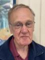 Older man with glasses, short hair, and a burgundy shirt, standing indoors near a painting or poster.
