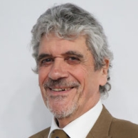 Smiling older man with grey curly hair and beard, wearing a tan suit and white shirt, against a plain background.