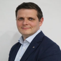 Man in a navy suit and light blue shirt smiling at the camera against a plain white background.