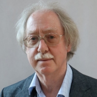 Older man with white hair, glasses, and moustache, wearing a grey suit jacket and light blue shirt.