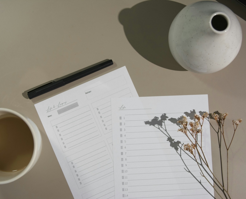 Two sheets of paper, a pen, a cup of coffee, a vase, and dried flowers are arranged on a table.